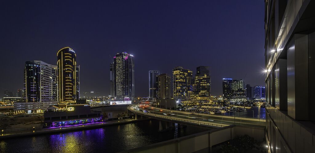 Une vue nocturne d’une ville, avec des gratte-ciel illuminés, un pont et une étendue d’eau reflétant les lumières.