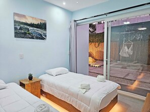 Un dormitorio con dos camas, una mesita de noche de madera con una planta y un ventanal con vistas a un jardín.