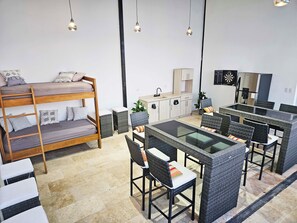 Una habitación moderna y bien iluminada con literas, una mesa de comedor con sillas de mimbre, una zona de cocina y un dardo.