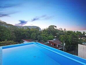 A rooftop pool with a view of a coastal area, surrounded by lush greenery and a mountain in the background.