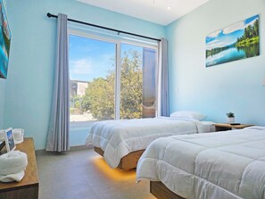 Hotellihuone, jossa on kaksi sänkyä, suuri ikkuna, jossa on verhot, seinällä taulu ja pieni pöytä, jonka päällä on kasvi.