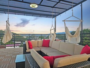 A covered outdoor seating area with a sofa and hanging chairs, overlooking a scenic landscape.