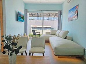 A modern living room with a sofa, chairs, a coffee table, and a vase of dried flowers. There is a large window with a view of a building and a mountain in the background.