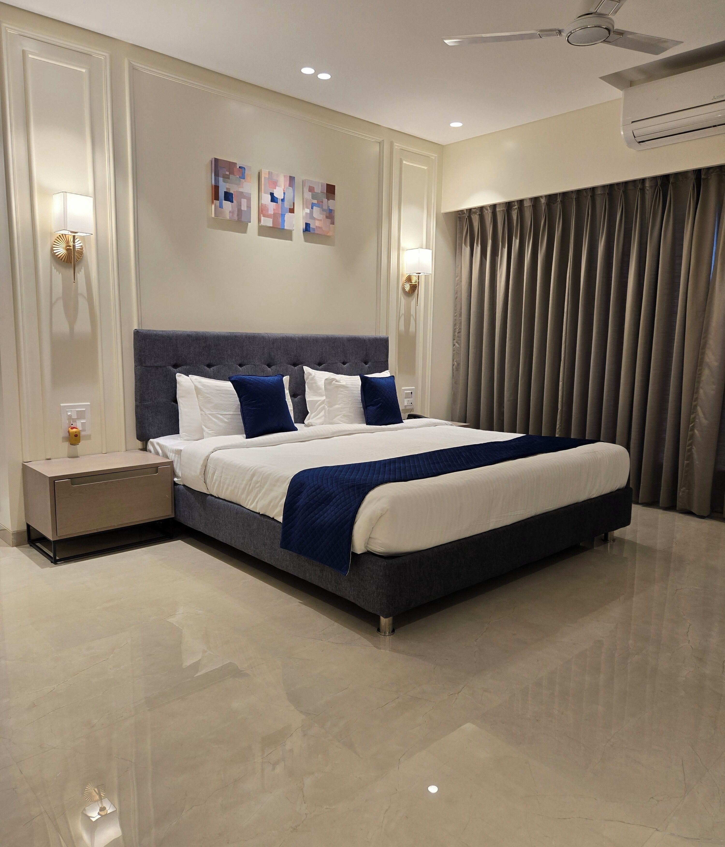 A modern hotel bedroom with a large bed, a nightstand, a wall-mounted lamp, and a ceiling fan.