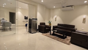 A modern living room with a glass partition, a piano, sofas, and a refrigerator. - Western Hotel (Chandrapur)