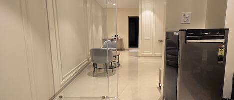 A modern hotel room with a glass partition, a refrigerator, and a small seating area with a table and chairs.