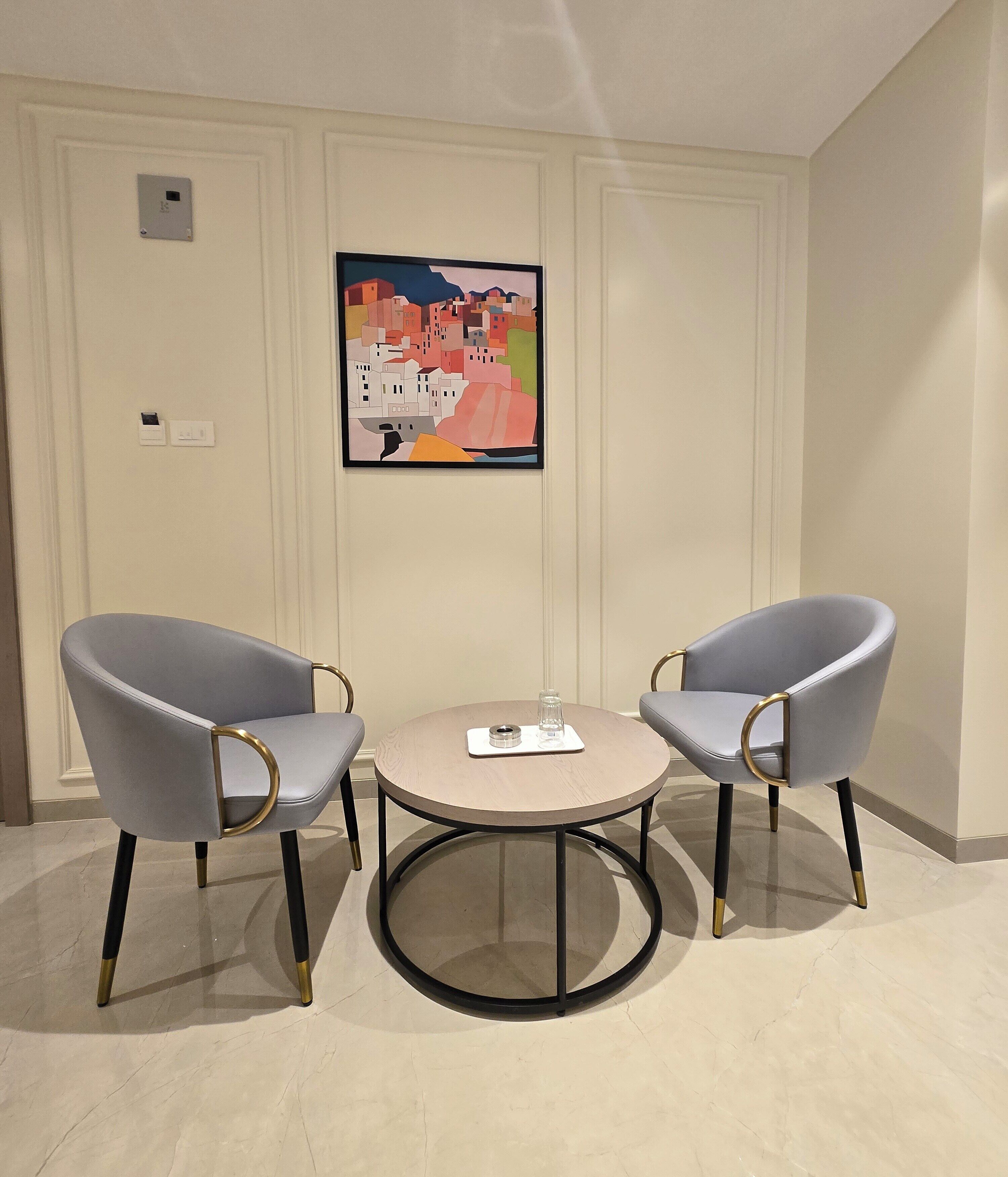 A room with two grey upholstered chairs with gold accents, a round table, and a colorful abstract painting on the wall.