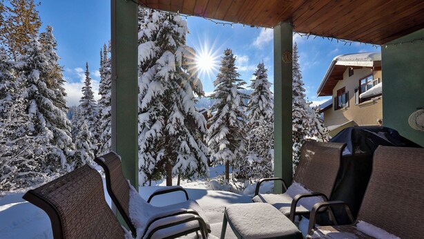 A covered outdoor seating area with snow-covered chairs and a view of snow-laden trees and a building in the background. - Upper floor 2 bedroom condo, great location close to village centre and next to ski runs (Sun Peaks)