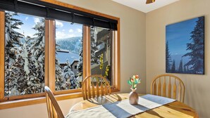 A dining area with a wooden table, chairs, a vase with flowers, a window with a view of snow-covered trees and a cabin, and a painting on the wall. - Upper floor 2 bedroom condo, great location close to village centre and next to ski runs (Sun Peaks)