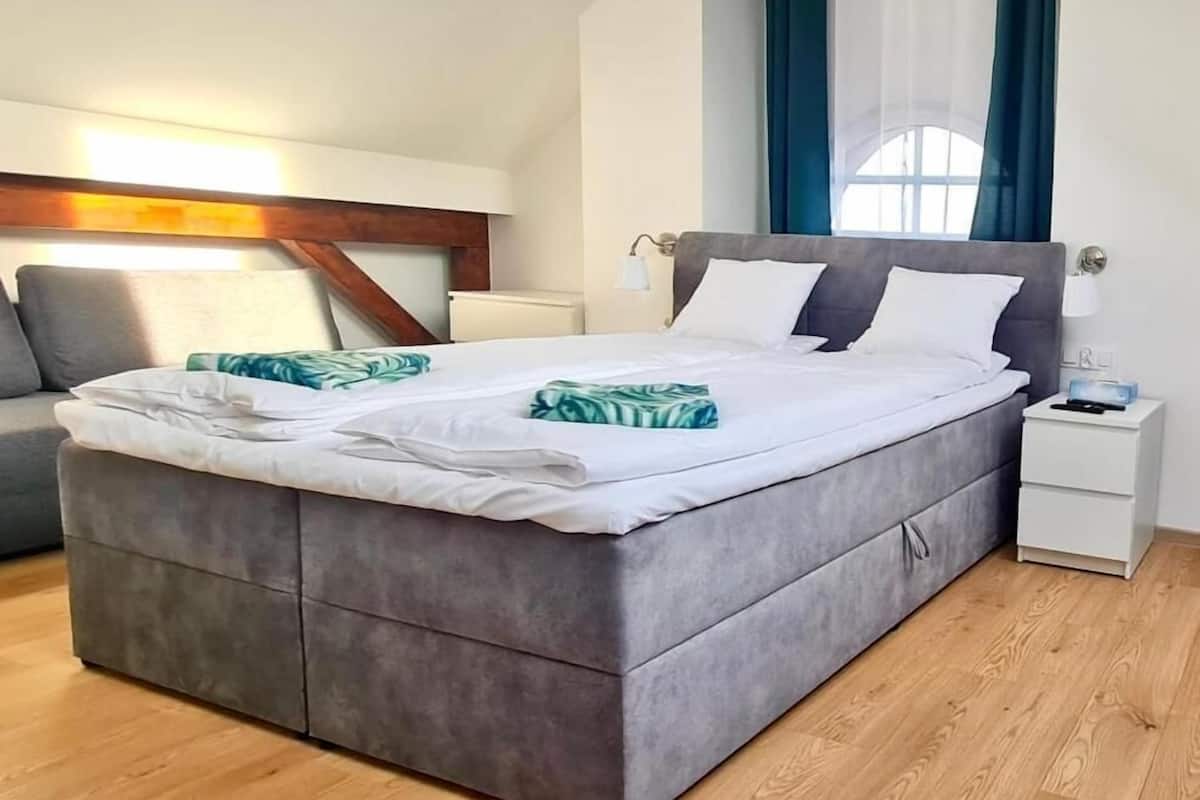 Un dormitorio moderno con una cama grande, una mesita de noche, una lámpara y vistas al exterior a través de una ventana con cortinas azules.