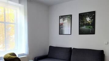 Una sala de estar con un sofá gris oscuro, un sillón azul y una mesita redonda. Hay dos cuadros enmarcados en la pared, una ventana con cortinas y una lámpara en el techo.