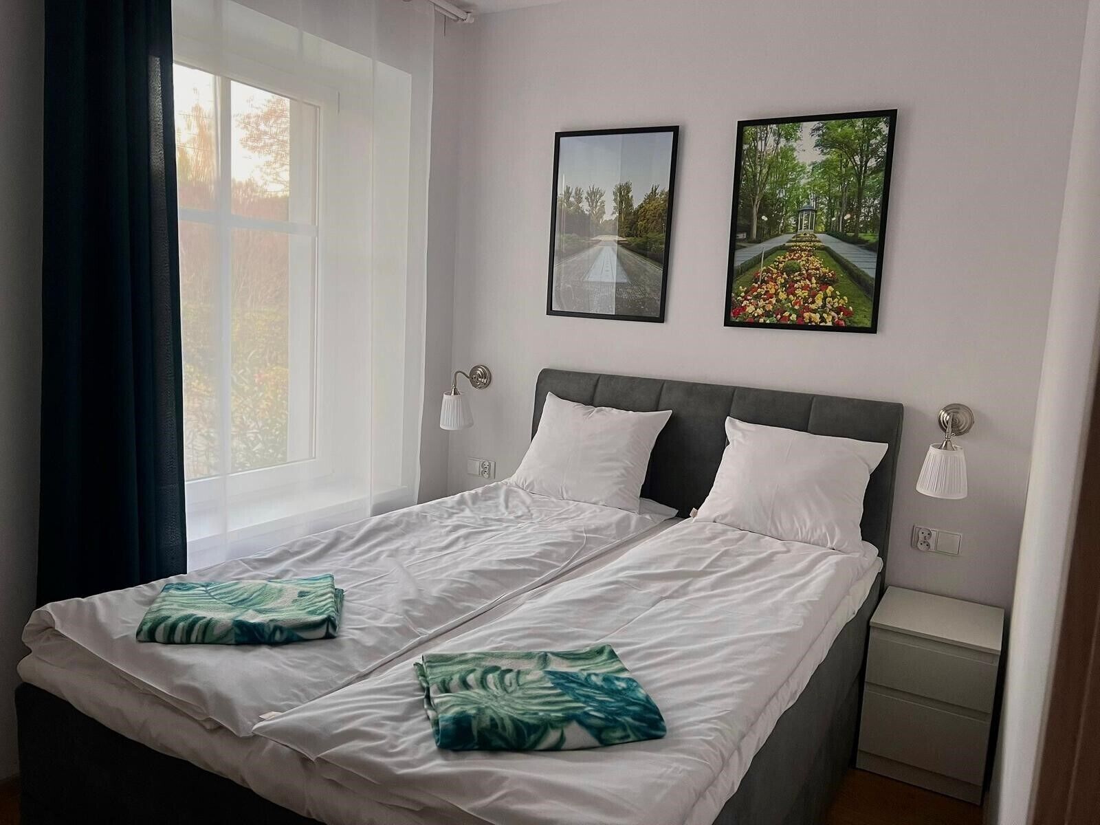 A double bed with white linens and two pillows, a bedside table with a lamp, and two framed pictures on the wall.