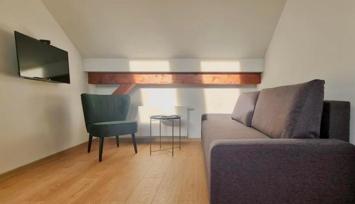 Un espacio habitable pequeño y moderno con techo de madera, un televisor de pantalla plana montado en la pared, un sillón verde, un sofá gris y una pequeña mesa redonda.