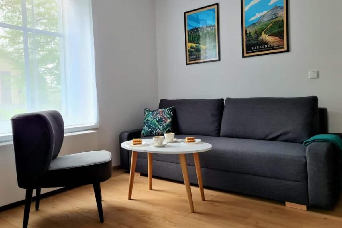 Una sala de estar moderna con un sofá gris, una silla negra y una pequeña mesa redonda con tazas y un libro. Dos cuadros enmarcados cuelgan de la pared.
