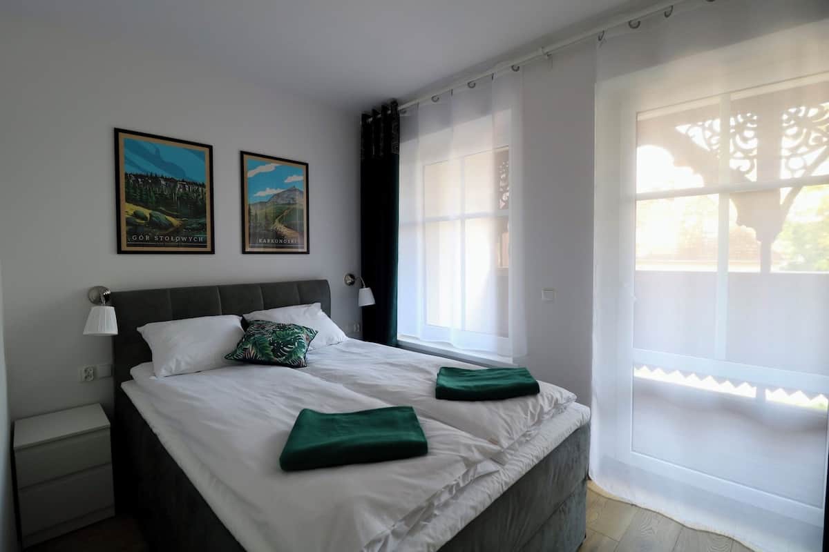 Un dormitorio con cama, mesita de noche, lámpara, dos cuadros enmarcados y una ventana con cortinas.