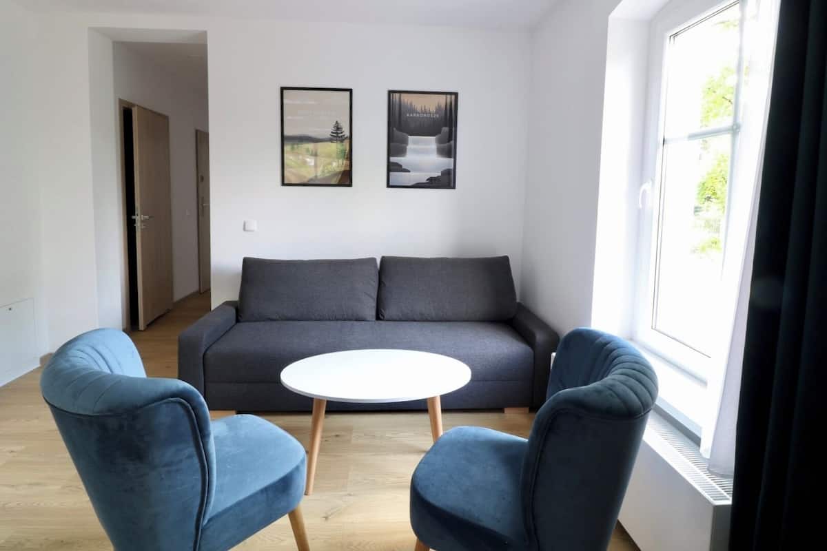 Una sala moderna con un sofá gris, dos sillones azules, una mesita blanca y dos cuadros enmarcados en la pared.