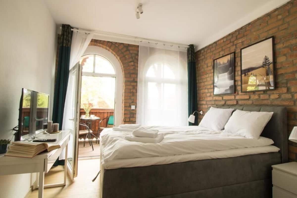 Un dormitorio con pared de ladrillo, una cama grande con ropa de cama blanca, un escritorio con una computadora portátil y vistas a una terraza exterior.