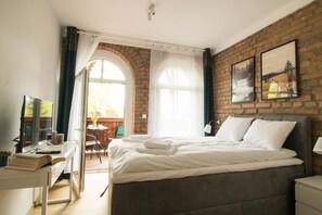 Un dormitorio con pared de ladrillo, una cama grande con ropa de cama blanca, un escritorio con una computadora portátil y vistas a una terraza exterior.