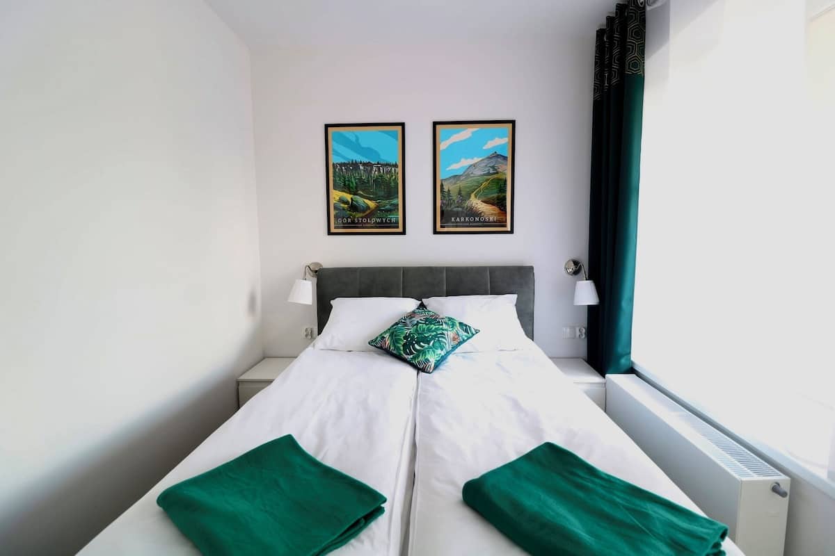 Una habitación de hotel con una cama, dos cuadros en la pared, una ventana con cortinas y una lámpara de noche.