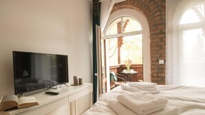 Un dormitorio con televisión sobre un tocador blanco, una cama con ropa de cama blanca y vistas a un balcón con mesa y sillas.