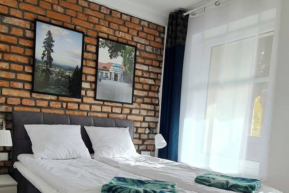 Un dormitorio moderno con una pared de ladrillo decorativa, una cama grande con ropa de cama blanca, dos almohadas verdes decorativas, una mesita de noche, una lámpara y una ventana con cortinas.