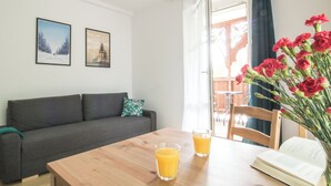 Un salón moderno con un sofá, una mesa de madera con dos vasos de jugo de naranja, un libro y un jarrón con flores rojas.