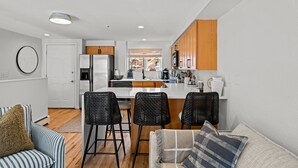 A modern kitchen with white cabinets, a central island, and black bar stools. There is a dining area with a striped sofa and a round mirror on the wall. - New Listing! Executive Rental - Steps to Vibrant Pearl Street Mall (Boulder)