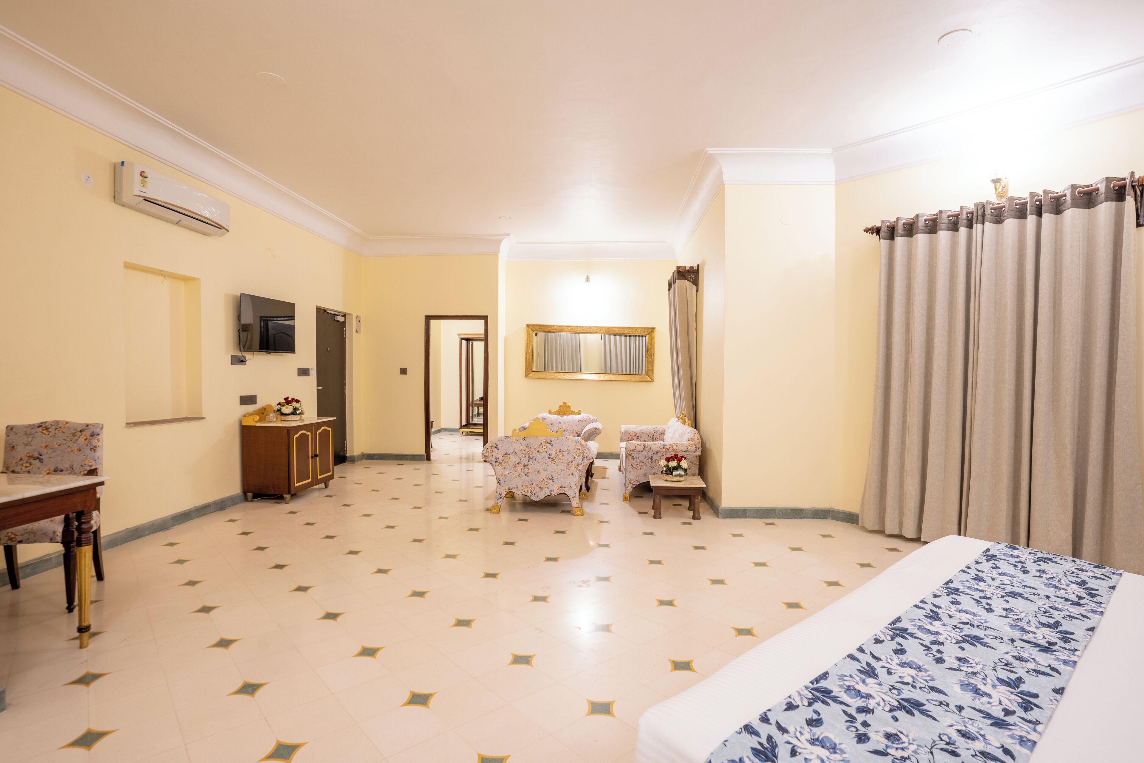 A hotel room with a bed, two armchairs, a small table, a TV, an air conditioning unit, and a door leading to another room.