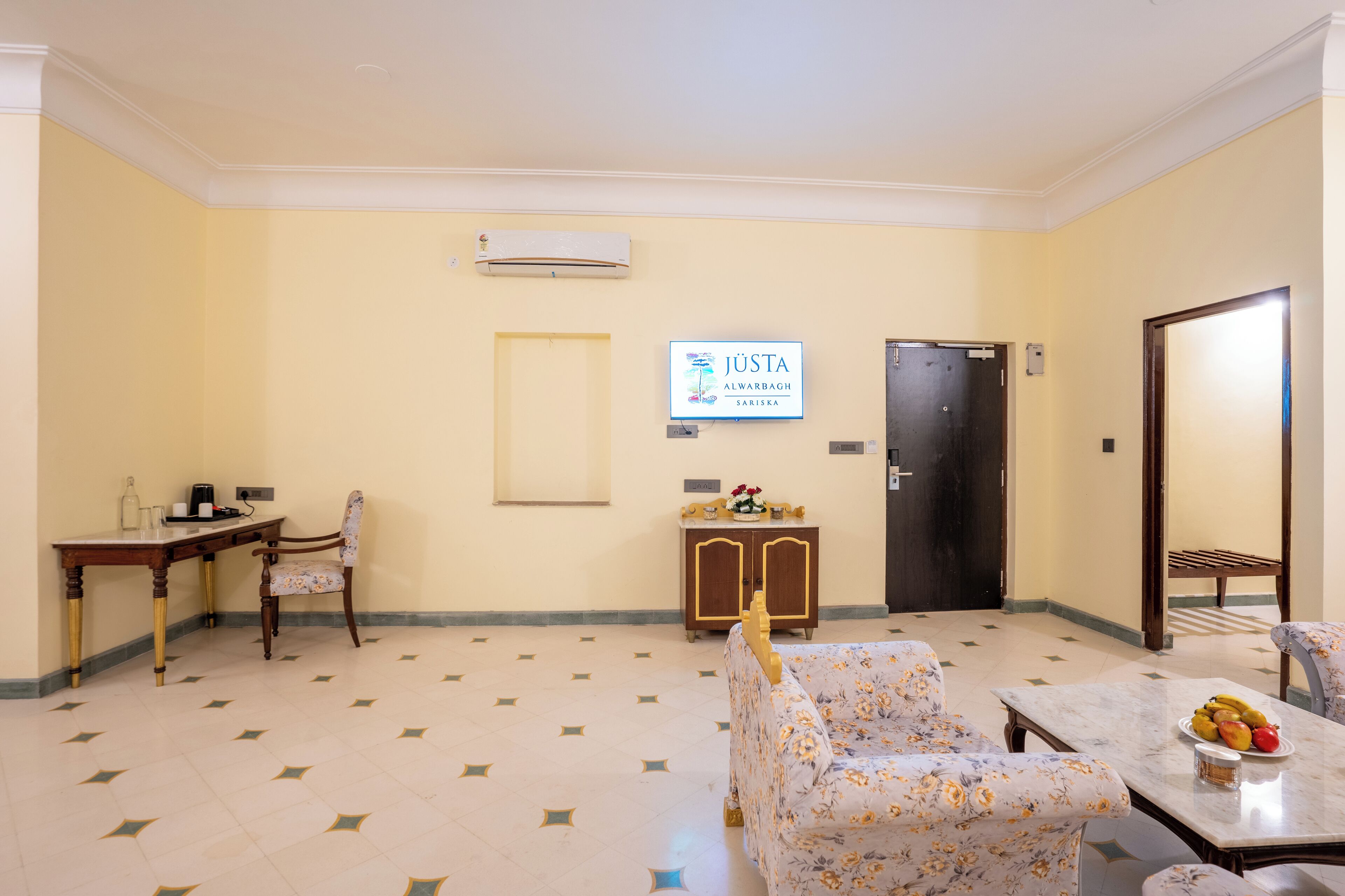 A hotel room with a dining area, a sofa, a table with fruit, a wooden cabinet, and a door.