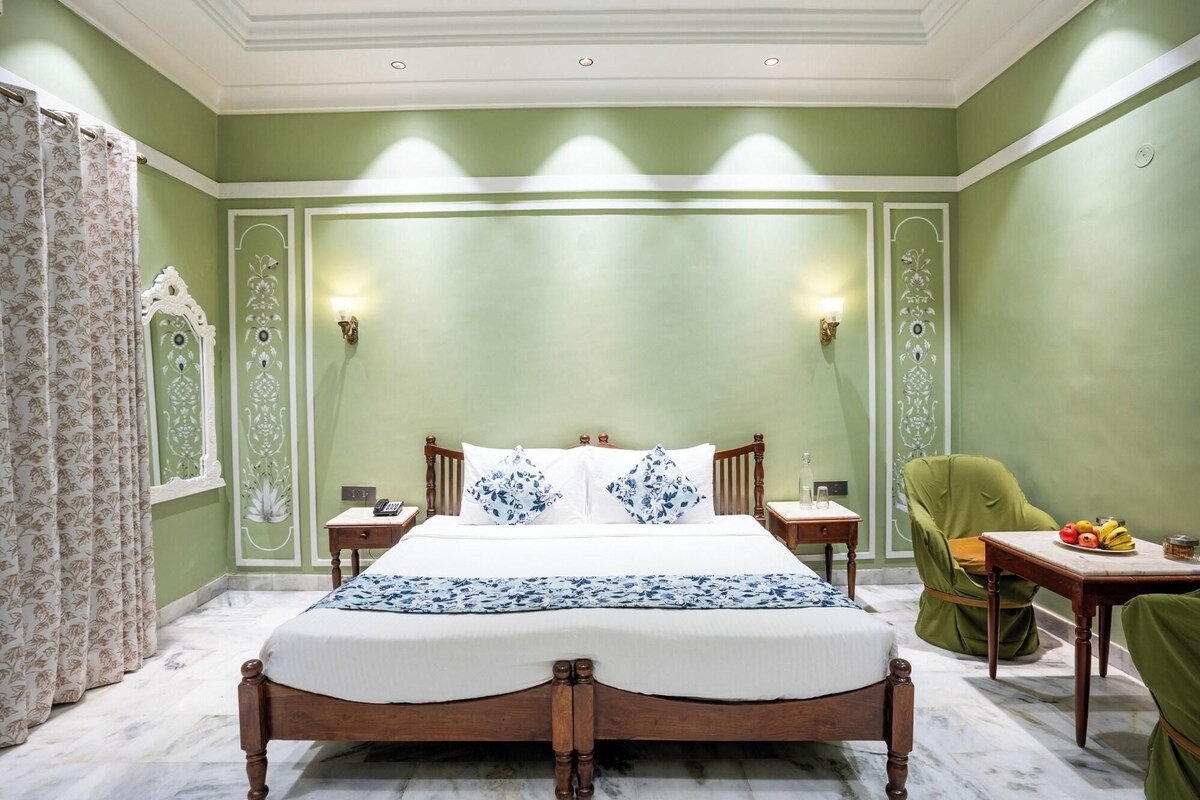 A hotel room with a large bed, two bedside tables, a green armchair, and a small table with fruit.