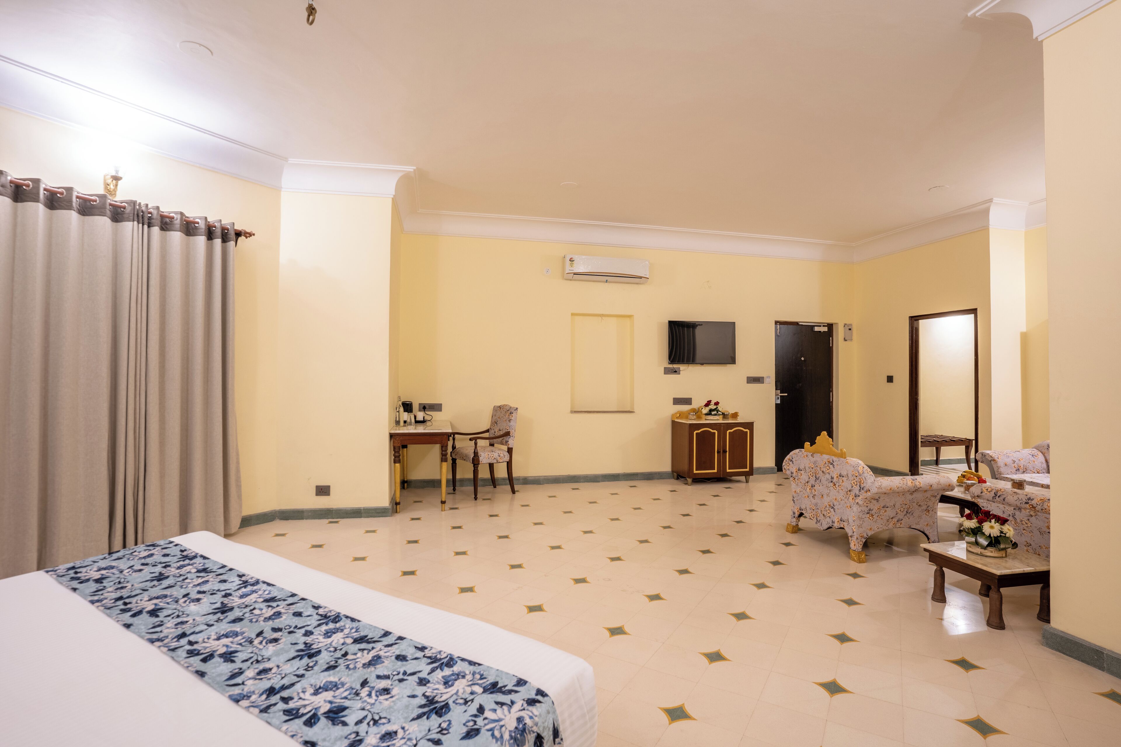 A hotel room with a bed, a desk, a chair, a sofa, a TV, and an air conditioning unit.