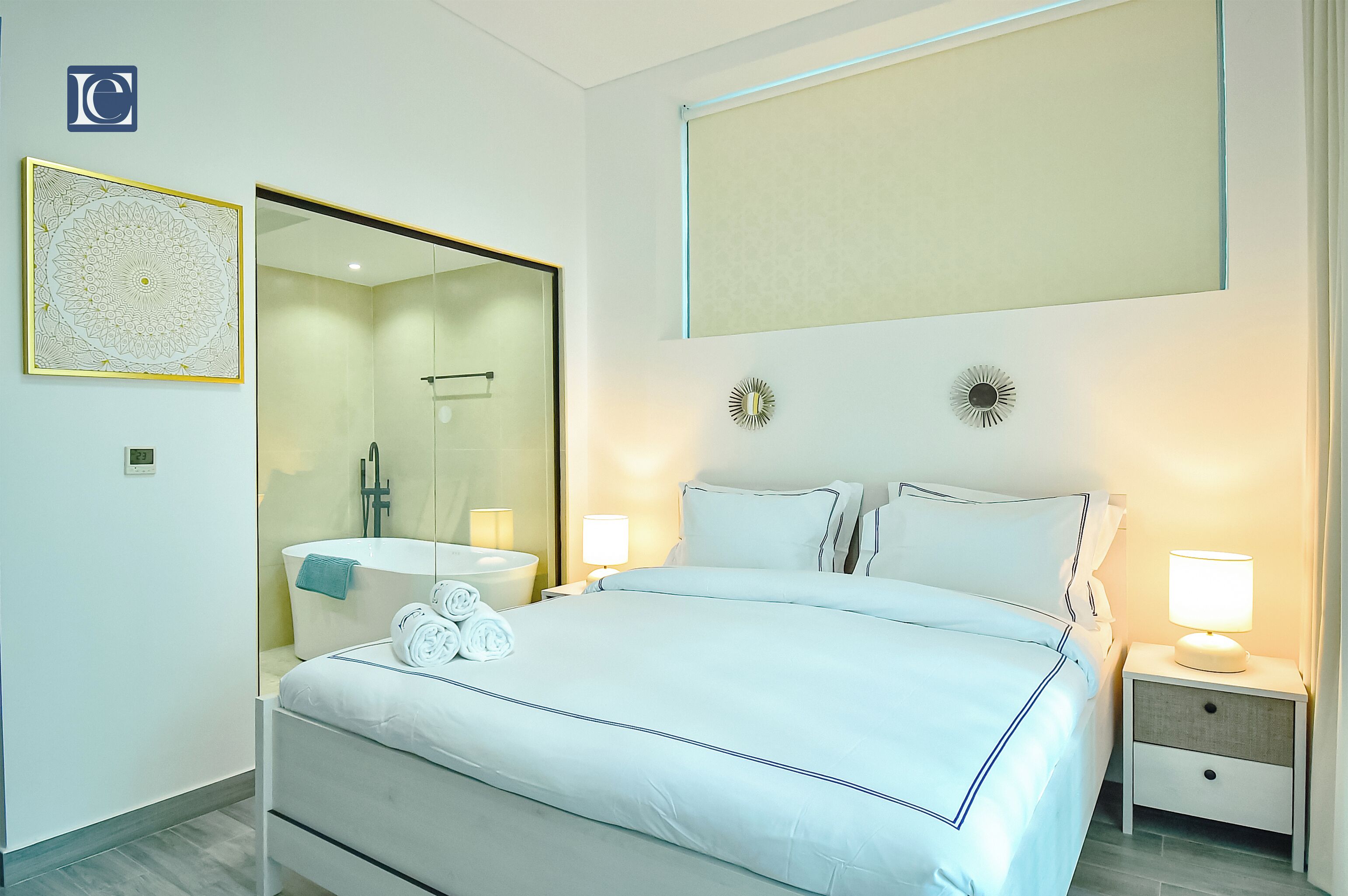 A modern hotel room with a large bed, a bathroom with a glass shower, and a nightstand with a lamp.