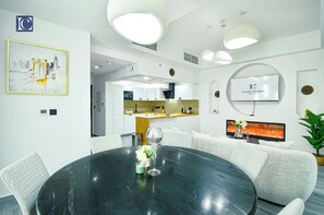A modern, well-lit dining area with a round table, white upholstered chairs, and a large flat-screen TV displaying 'Luxury Escapes'.