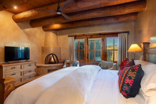 A bedroom with a large bed, wooden ceiling beams, a television on a dresser, and a window with curtains.