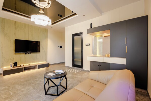 A modern living room with a flat-screen TV, a sofa, a coffee table, and a built-in kitchen counter. - CVR'S HOTEL GLASS BIRD (Ravulapalem)