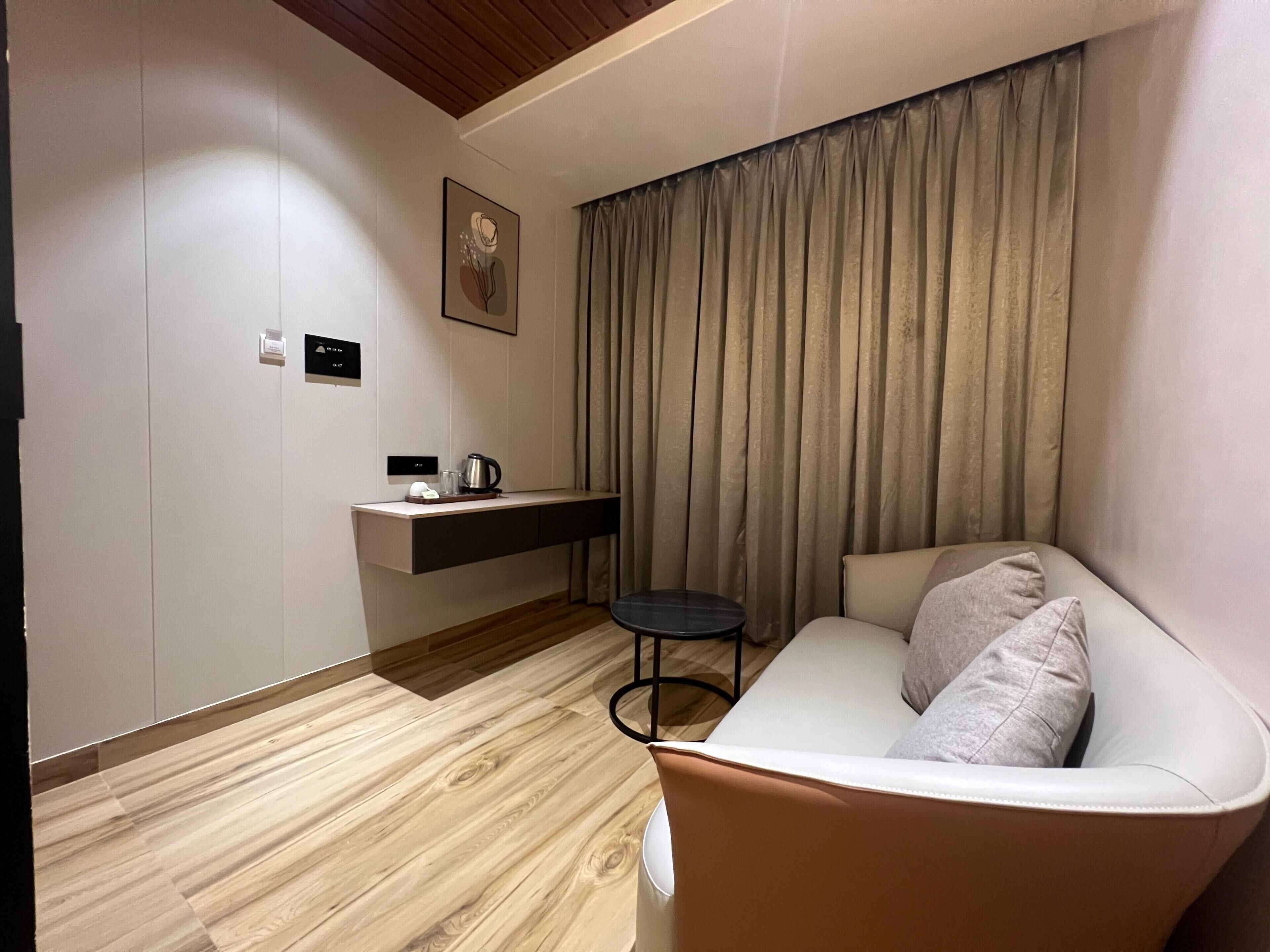 A modern hotel room with a wooden floor, a sofa with cushions, a small table, a wall-mounted charging station, and a window with curtains.