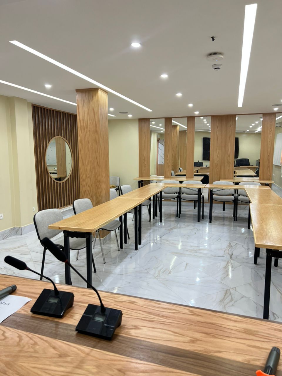 Un salón de conferencias con largas mesas, sillas y un panel de madera con espejo.
