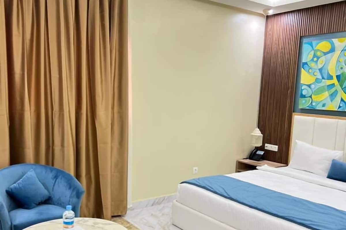 A hotel room with a bed, two blue armchairs, a small round table with a water bottle, a nightstand with a lamp, and a painting on the wall.
