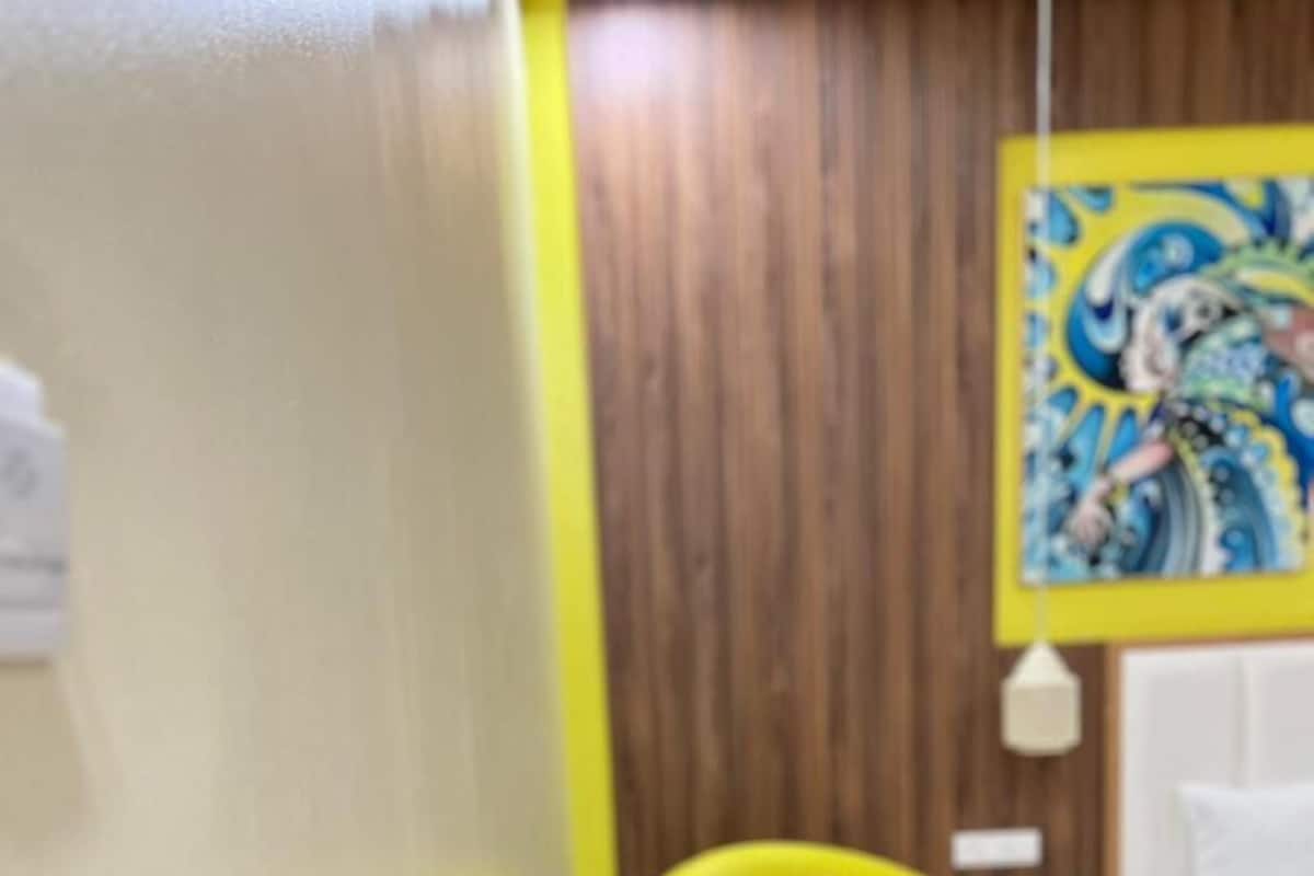 A hotel room with a yellow armchair, a small round table with water bottles, a white sofa, and a wooden wall with a colorful painting.