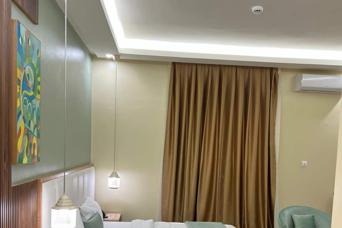 A modern hotel room with a bed, bedside tables, a small desk, a chair, and a painting on the wall.
