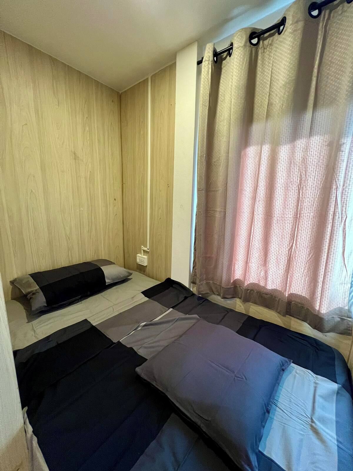 Een kleine, studioachtige accommodatie met een houten wand, een bed met een grijs en zwart matras, en een raam met een dunne gordijn.