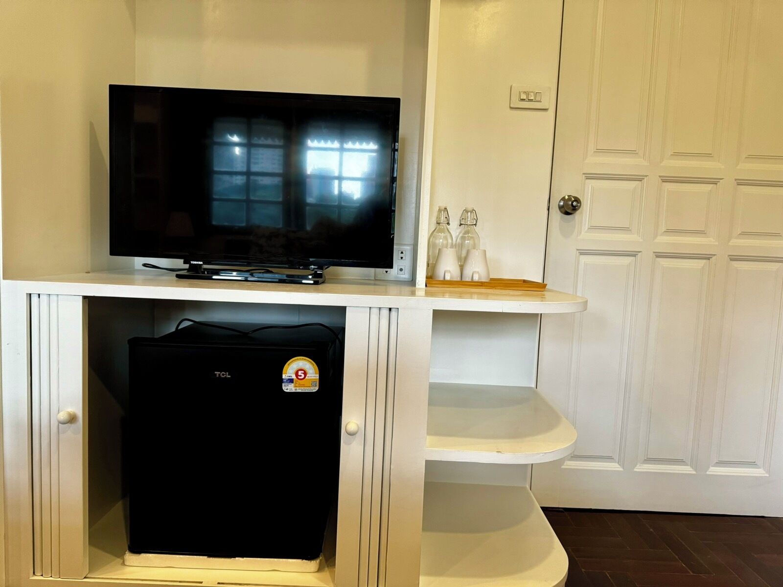A television on a white cabinet with a black device below it, a white door with a handle, and a shelf with two bottles and a tray.
