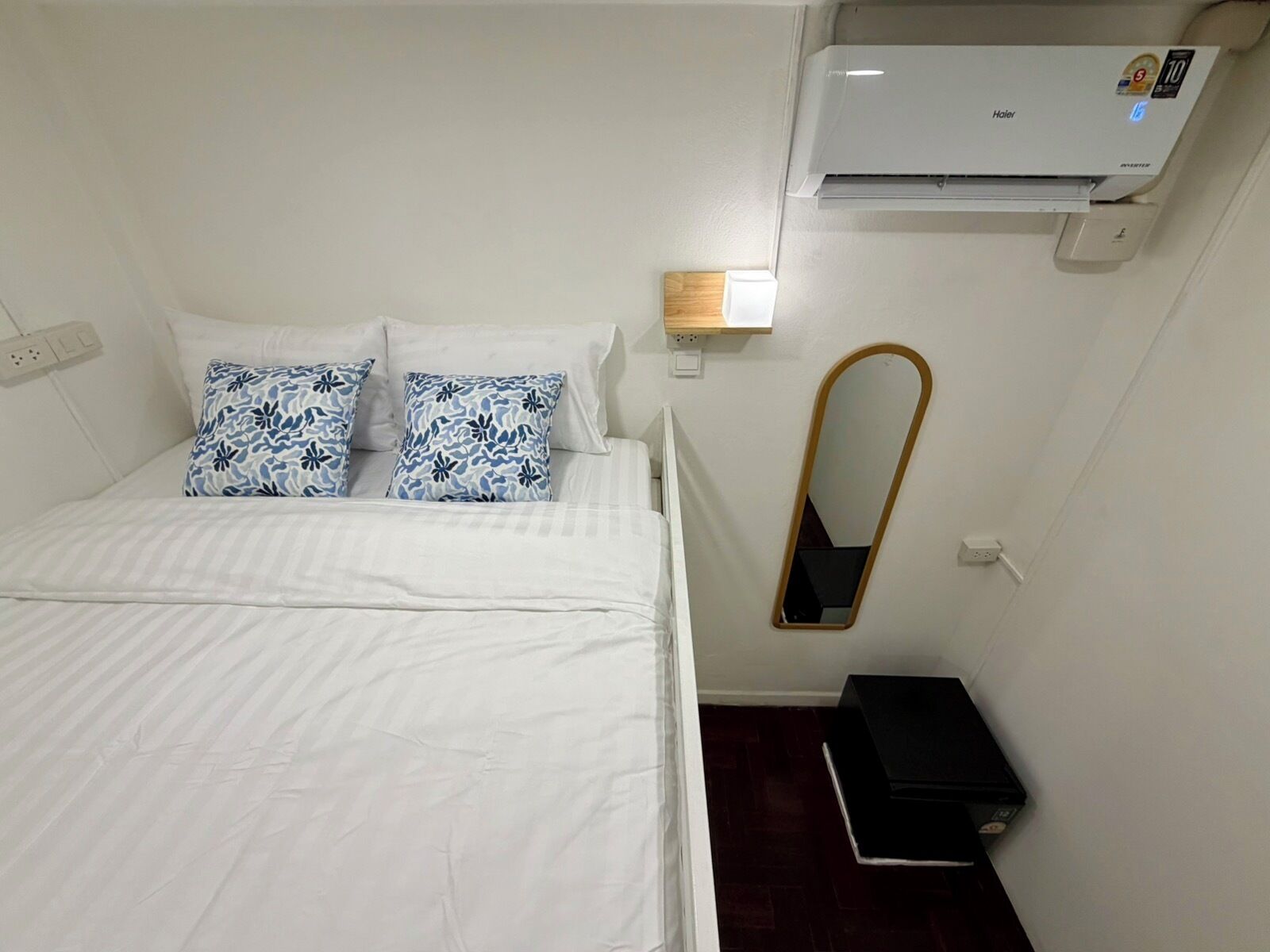 Ein Hotelzimmer mit Bett, Wandklimaanlage, Nachttisch mit Lampe und einem kleinen schwarzen Gegenstand auf dem Boden.