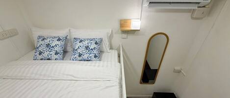 Ein Hotelzimmer mit Bett, Wandklimaanlage, Nachttisch mit Lampe und einem kleinen schwarzen Gegenstand auf dem Boden.