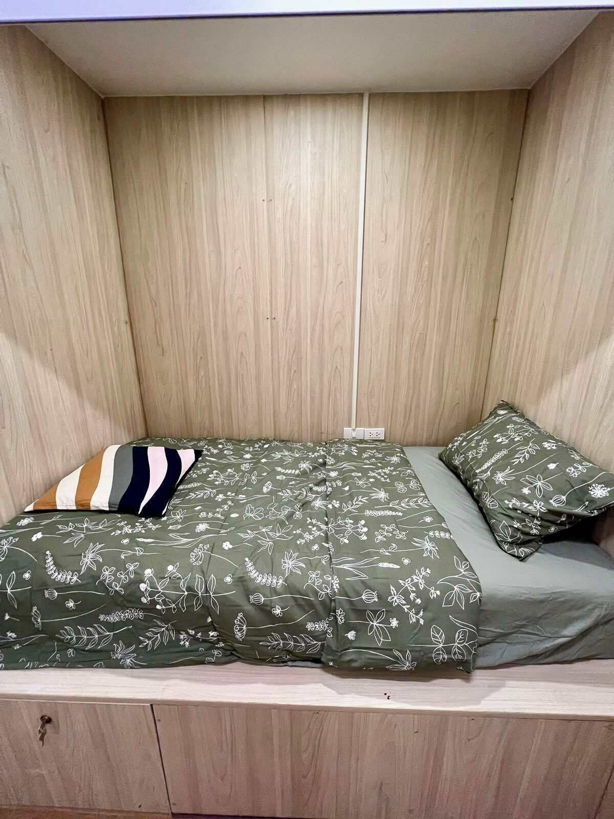 Una cama pequeña con estructura de madera y colcha estampada con motivos florales verdes, junto con almohadones a juego.