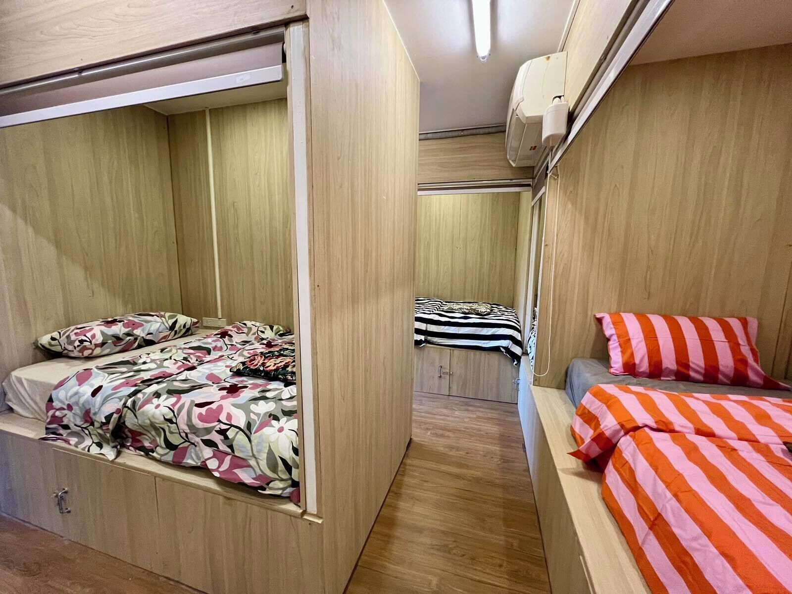 A compact room with two beds, one with floral bedding and the other with striped bedding. There is a wooden wardrobe and a small table.