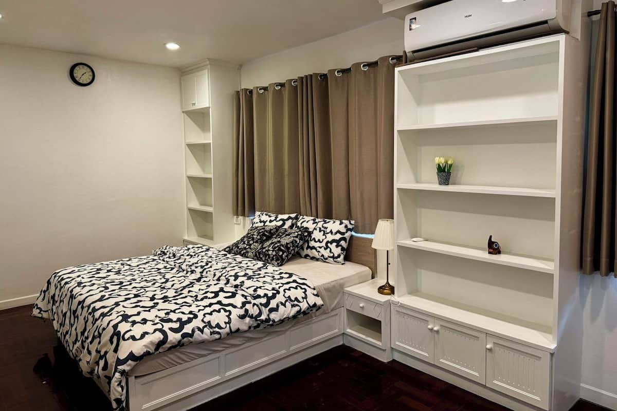 Un dormitorio con cama, mesita de noche, lámpara, estantería y aire acondicionado.