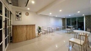 A spacious, well-lit room with a wooden reception desk, multiple tables and chairs, and a large window providing natural light. - Comfy Crib Hostel 2  (Bangkok)