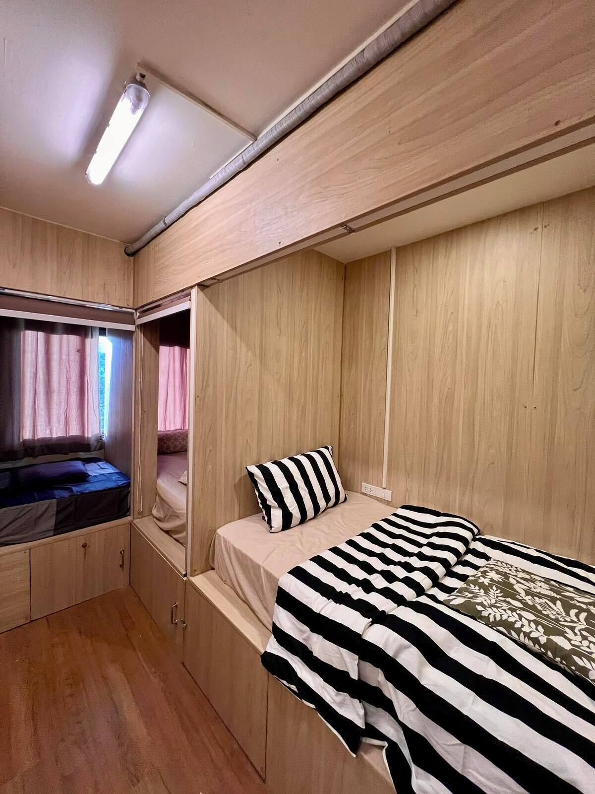 A compact bedroom with a bunk bed, a single bed with striped bedding, a wooden wardrobe, and a window with curtains.