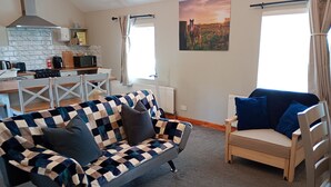 A living room with a checkered sofa, a blue armchair, a kitchen in the background, and a painting on the wall. - Mattys Pub & Apartments (Muine Bheag)